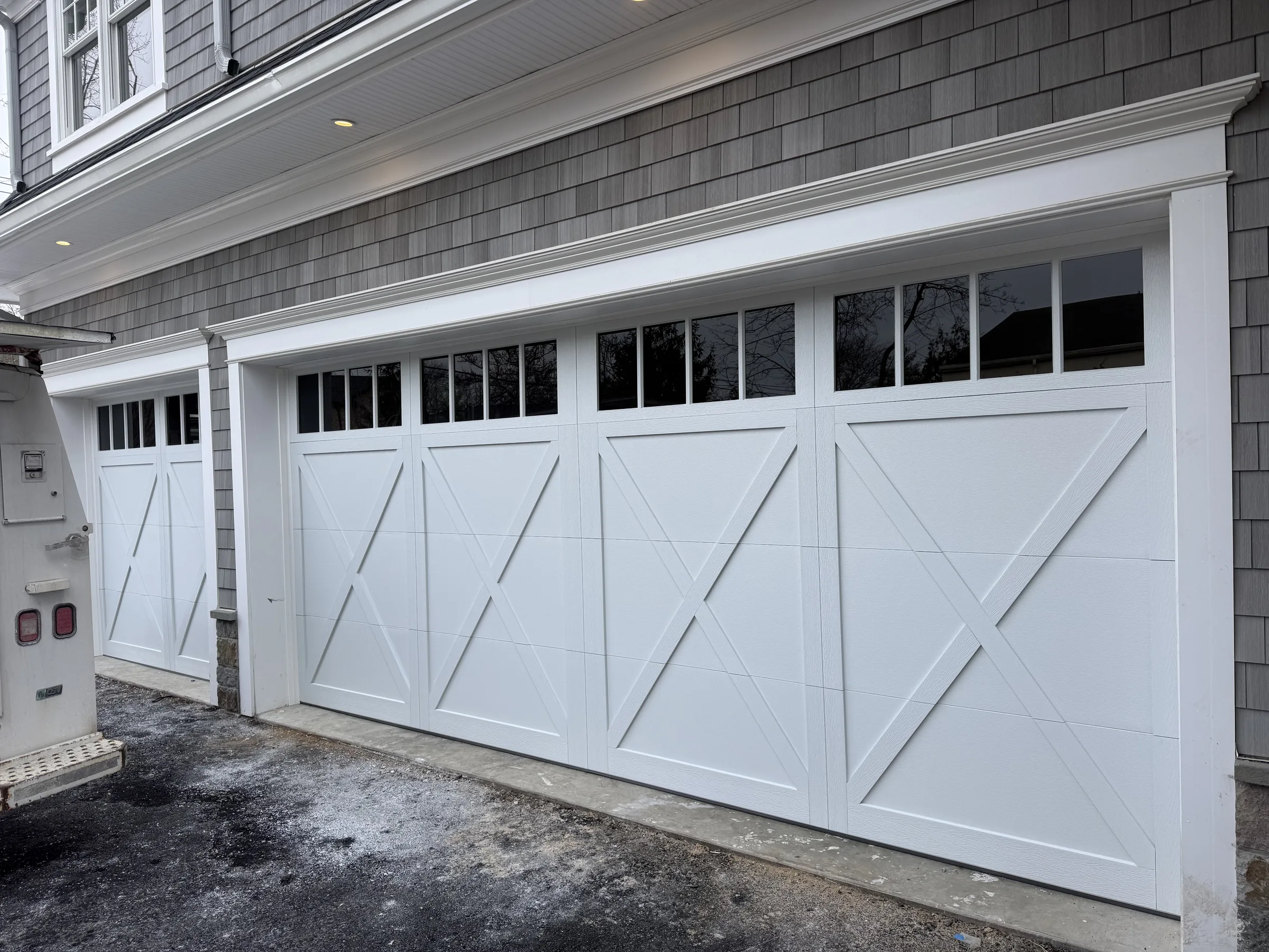 White carriage-style garage doors with X-brace panel detailing and upper windows on a gray-shingled residential home.